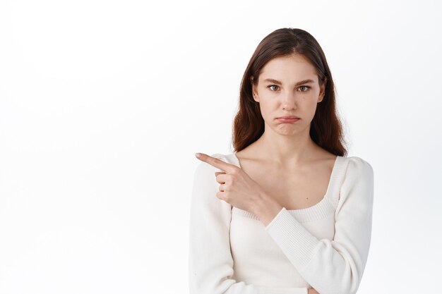 Displeased young woman pointing index finger aside, showing bad promo, sulking upset, looking disappointed, standing against white wall