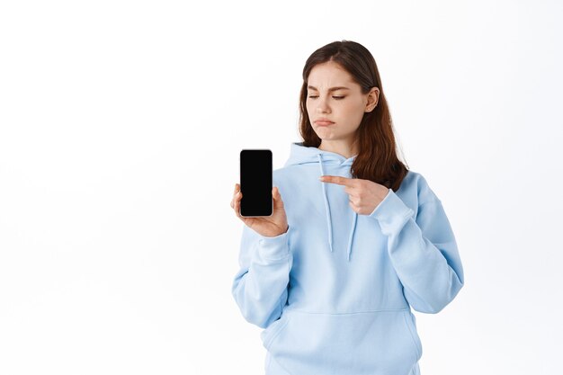 Displeased young woman showing bad application on her phone, pointing at empty smartphone screen and grimacing upset, standing against white wall