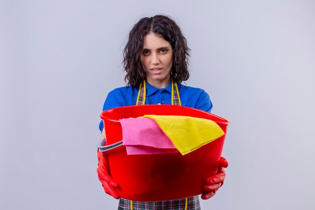 Free photo displeased young woman wearing apron and rubber gloves holding bucket with cleaning tools with serious face over white wall