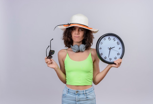 Foto gratuita una giovane donna scontenta con i capelli corti in top corto verde che indossa cappello da sole con occhiali da sole e orologio da parete su uno sfondo bianco