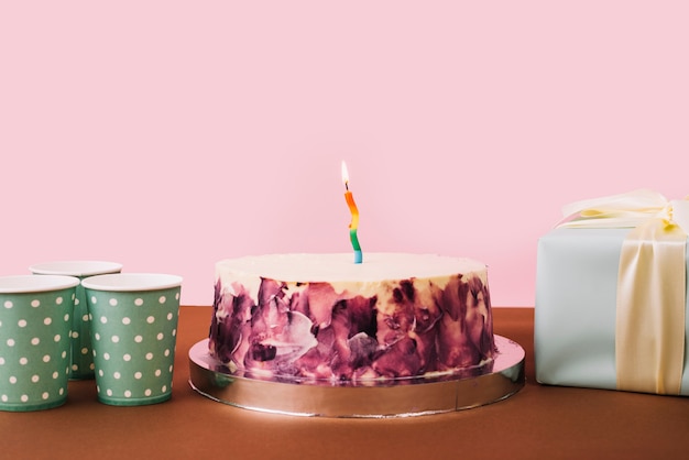 Disposable glasses; gift box and tasty cake on desk against pink backdrop