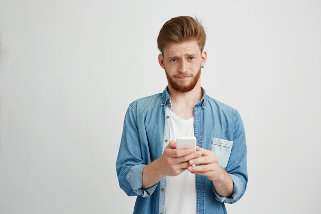 Dissapointed upset young man with beard holding smart phone looking at camera.