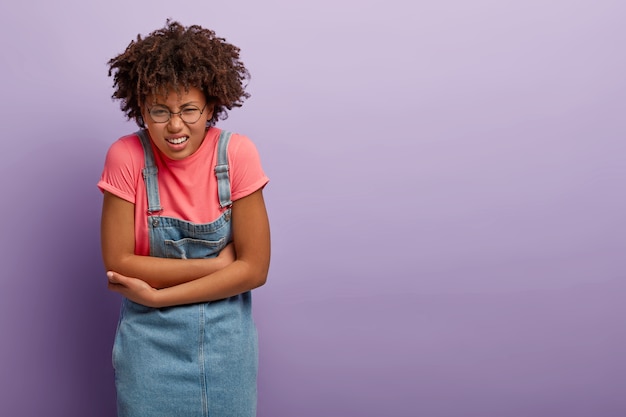 La donna afroamericana insoddisfatta tiene le mani sulla pancia, stringe i denti per sentimenti spiacevoli, ha disturbi o crampi mestruali, sente fastidio alla pancia, indossa denim sarafan.