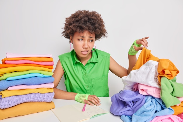 Dissatisfied African American woman picks dirty clothes from pile works in laundry makes notes in notebook poses against white wall feels aversion has disgusting face. Big laundering concept