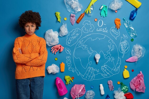 Dissatisfied Afro American woman activist engaged in antipollution campaign, looks angrily at camera with hands crossed, poses against blue background