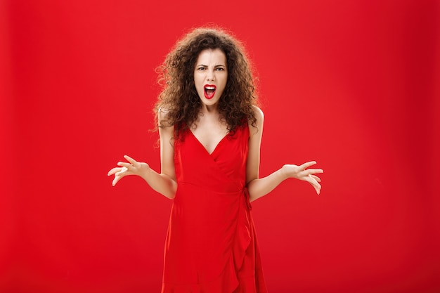 Dissatisfied angry and pissed attractive curlyhaired female in stylish red dress arguing and frownin...