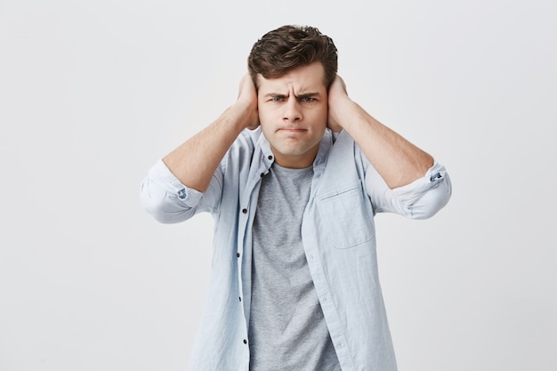 Dissatisfied and angry young caucasian male dressed in light blue shirt, frowning his face, closing ears with palms, annoyed and tired of noise, refusing to listen to someone`s words. Body language