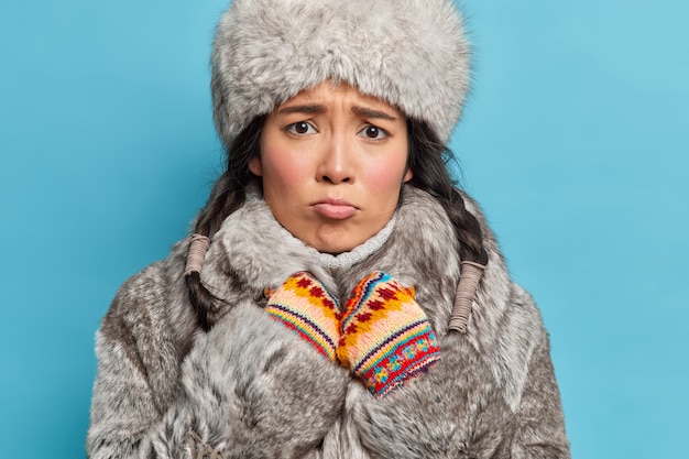 Бесплатное фото Недовольная азиатская женщина живет на крайнем севере, одетая в серую верхнюю одежду, носит теплые перчатки, хмурится и дрожит от холода, изолировавшись от синей стены