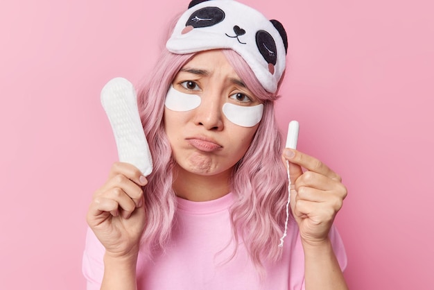 Dissatisfied Asian woman purses lips wants to cry has bad mood holds sanitary napkin and tampon tilts head wears sleepmask casual t shirt isolated over pink background Absorbency and hygiene