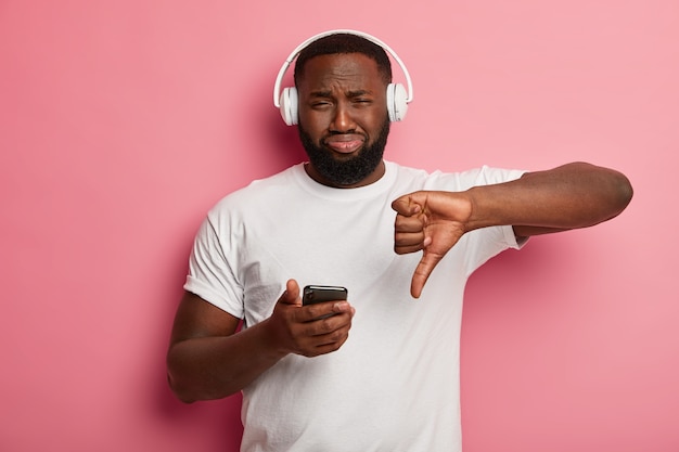 Dissatisfied bearded black man shows dislike gesture, doesn't like song from playlist, hears sound track in headphones