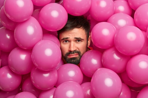 Недовольный бородатый европейец, уставший после вечеринки по случаю дня рождения, позирует вокруг множества розовых воздушных шаров