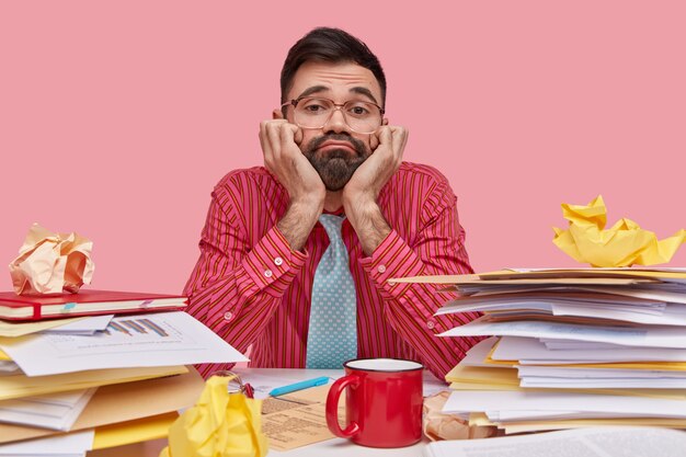 Dissatisfied bearded man keeps hands under chin, looks through spectacles with apathy, wears formal clothes, surrounded with stack of papers