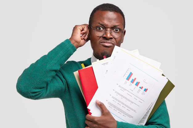Недовольный темнокожий мужчина составляет бухгалтерский отчет, держит бумаги с инфографикой, носит очки для хорошего зрения.