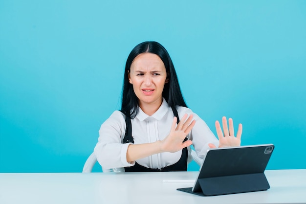 Dissatisfied blogger girl is showing stop gesture with hands by looking at tablet camera on blue background