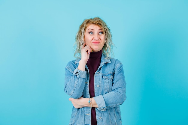 Dissatisfied blonde woman is looking away by holding hand on chin on blue background