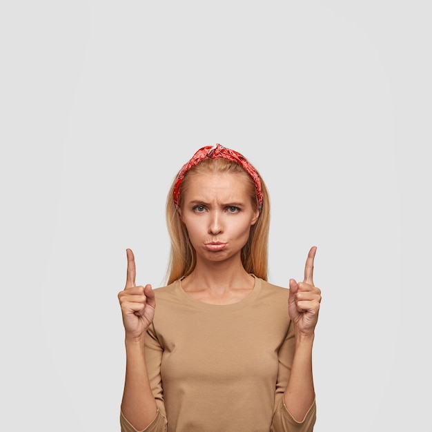 Free photo dissatisfied blonde woman with offensive look, gloomy expression, points with both index fingers upwards