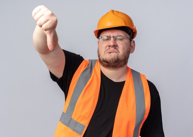 Недовольный строитель в строительном жилете и защитном шлеме смотрит в камеру с нахмуренным лицом, показывая пальцы вниз, стоя на белом фоне