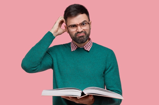 Dissatisfied Caucasian man with thick bristle, scratches head in bewilderment, wears spectacles and green sweater