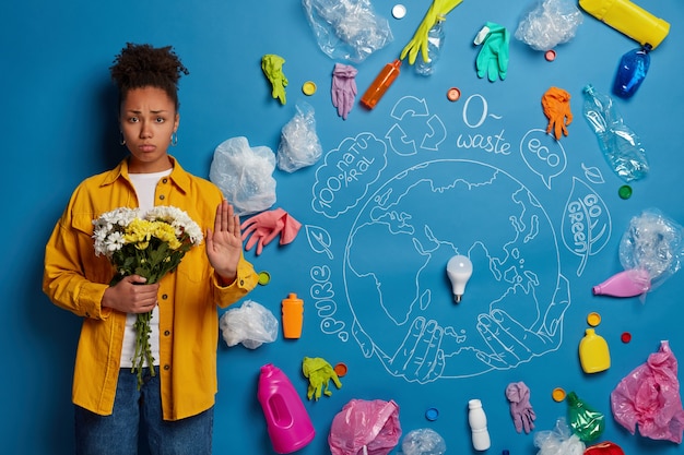 Dissatisfied curly haired afro woman makes stop gesture, holds flowers in hands, asks humanity to stop and think about cleaning nature