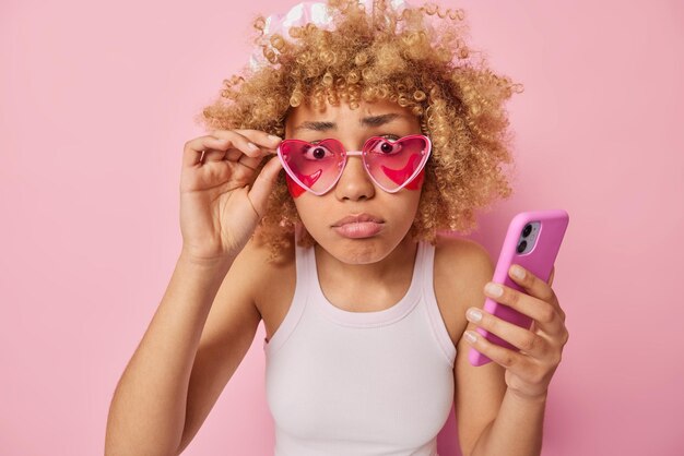 Недовольная кудрявая европейская женщина смотрит в камеру через розовые солнцезащитные очки, не может поверить своим глазам, наносит косметические повязки под глазами, держит мобильный телефон, одетый в футболку, позирует в помещении