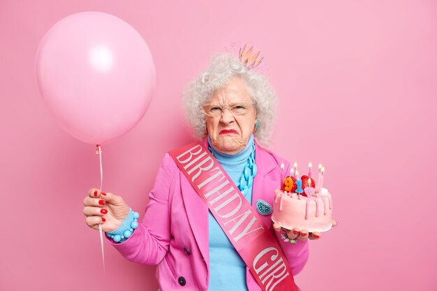 Dissatisfied curly haired senior lady celebrates birthday wears fashionbale clothes and jewelry poses with inflated balloon tasty cake looks with grumpy expression