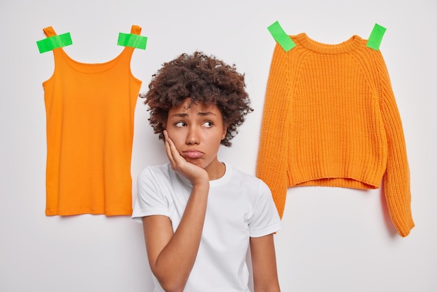 Dissatisfied curly haired woman looks away with sad thoughtful expression keeps hand on cheek dressed casually poses against white background with orange clothes plastered. Negative emotions