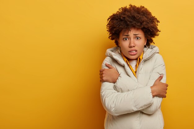 Dissatisfied dark skinned woman feels frozen after outdoor walk during frosty weather, wears warm white coat