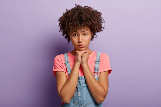 Dissatisfied dark skinned woman with curly hair, holds hands under chin, feels lonely and dejected after quarrel with boyfriend, has Afro hairstyle, wears casual t shirt isolated on purple