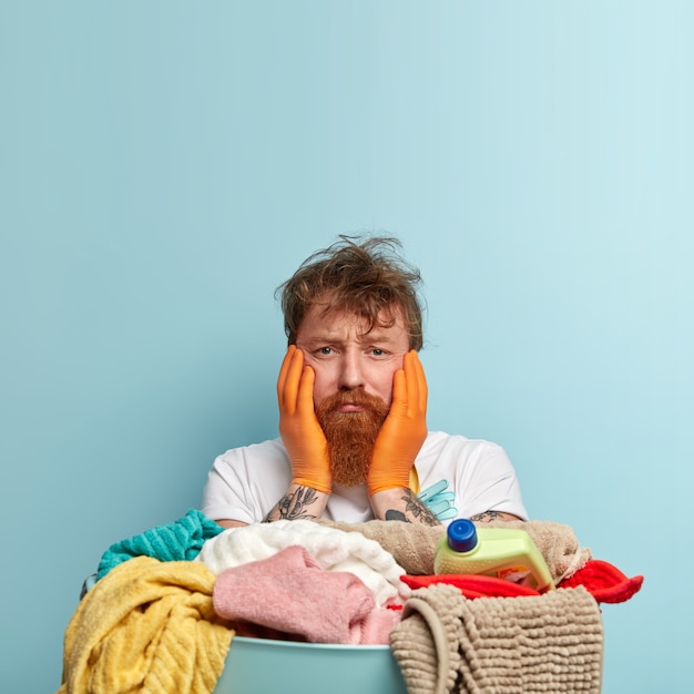 Dissatisfied desperate ginger man with messy hair, touches cheeks with both hands, overworked, has pile of dirty towels, stands over blue wall, blank space for your advertising content