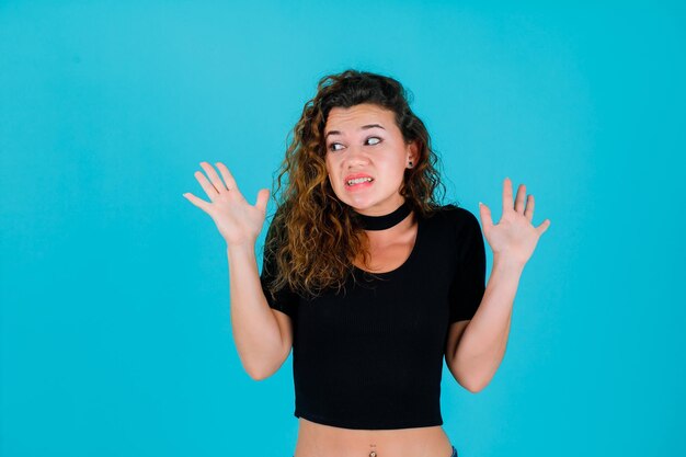 Dissatisfied girl is looking left by opening wide her hands on blue background