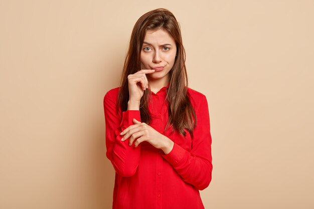 Dissatisfied good looking woman raises eyebrows, purses lips, looks angrily, keeps hand near mouth, wears red stylish shirt, stands over beige wall, discontent with somebodys suggestion