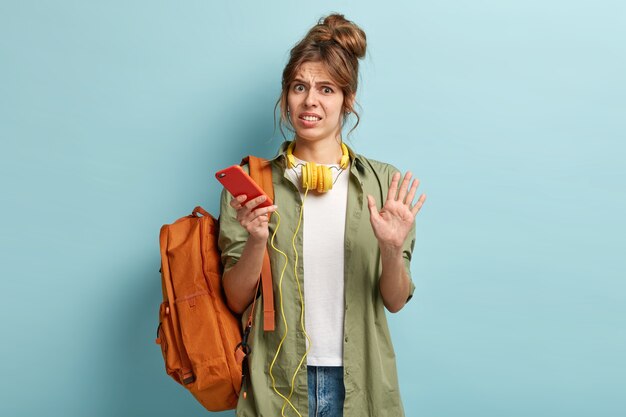 Dissatisfied hipster girl makes stop gesture in displeasure, listens music via modern cell phone and stereo headphones