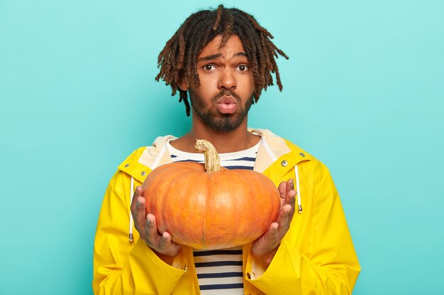 Dissatisfied hipster looks in displeasure, holds orange pumpkin, wears yellow raincoat, carries squash