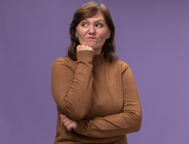 Недовольная женщина средних лет в коричневой водолазке смотрит в сторону, положив руку на подбородок и думает, стоя у фиолетовой стены