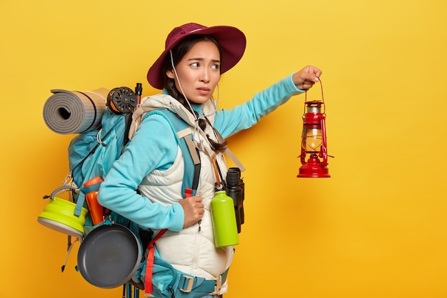 Dissatisfied mixed race female backpacker wears stylish hat and warm vest, holds kerosene lamp to explore surroundings in darkness