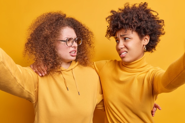 Le donne insoddisfatte di razza mista fanno un sorrisetto e si guardano infelici l'un l'altro si abbracciano e fanno selfie hanno qualche problema indossare abiti casual isolati su un muro giallo. reazioni umane negative