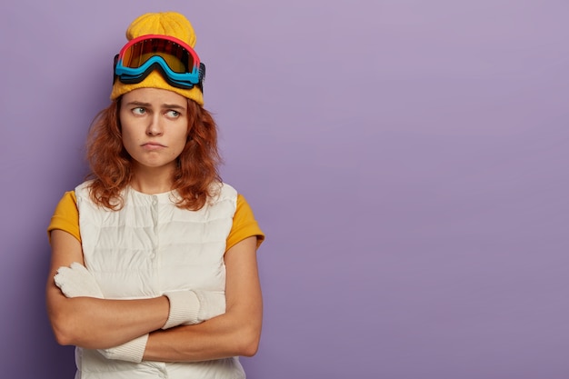 Dissatisfied redhaired woman keeps hands crossed, frowns face, being in bad mood, wears ski goggles and white vest, discontent , isolated over purple background.