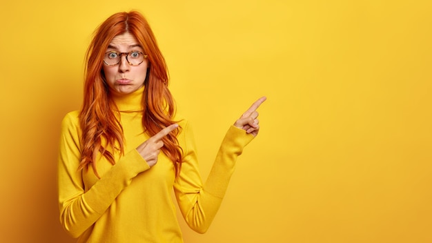 Free photo dissatisfied redhead freckled woman purses lower lip and points away on blank space shows something bad wears casual turtleneck spectacles.