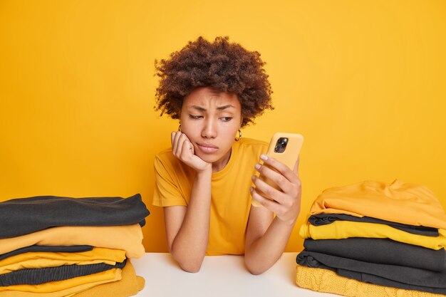 Dissatisfied sad woman feels tired after folding clothes looks attentively at smartphone checks newsfeed leans at table surrounded by two stacks of yellow and black folded laundry poses indoor