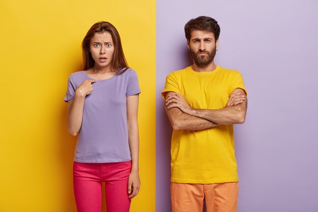 Dissatisfied sullen unshaven guy keeps arms folded, indignant young woman points at herself