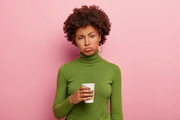 Dissatisfied tired African American woman holds takeaway coffee, tries to refresh after hard work, wears green polo neck sweater, sighs from tiredness, feels overworked