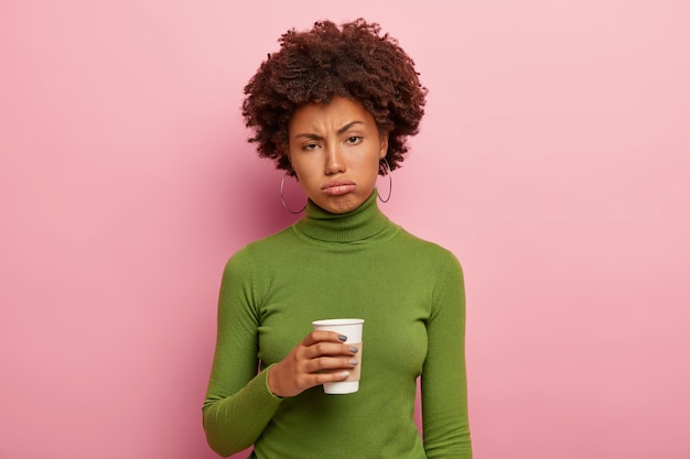 不満の疲れたアフリカ系アメリカ人の女性は、持ち帰り用のコーヒーを持って、ハードワークの後にリフレッシュしようとし、緑のタートルネックのセーターを着て、疲れからため息をつき、過労を感じます