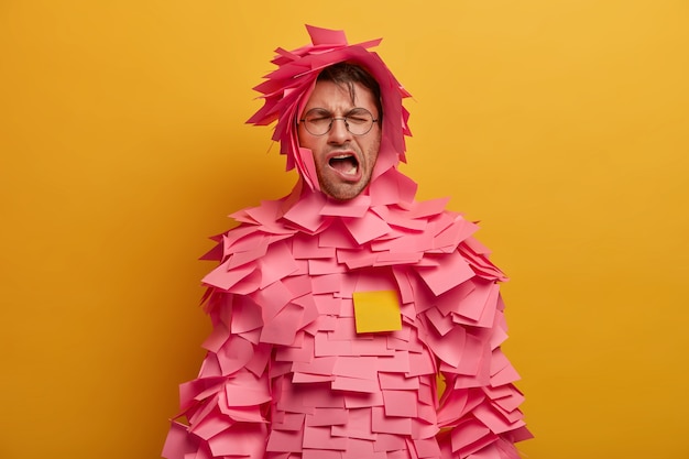 Free photo dissatisfied tired man yawns, opens mouth and keeps eyes shut, wears sticky notes outfit, has fun or foolishes around, poses over bright yellow wall. guy covered with stickers on body and over head
