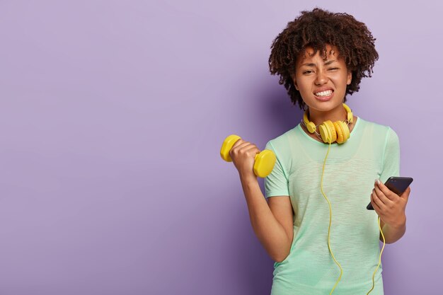 Dissatisfied tired woman dressed in sports clothing, raises hands with kettlebell, smirks face, holds cellphone connected to headphones