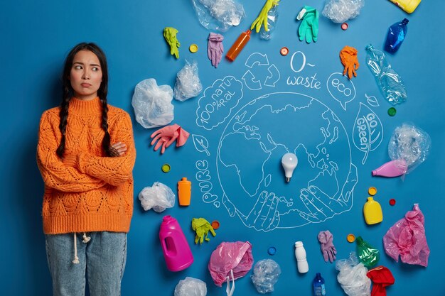 dissatisfied young Asian woman keeps arms folded, feels unhappy and concerned about environmental or nature problem, wears knitted orange sweater