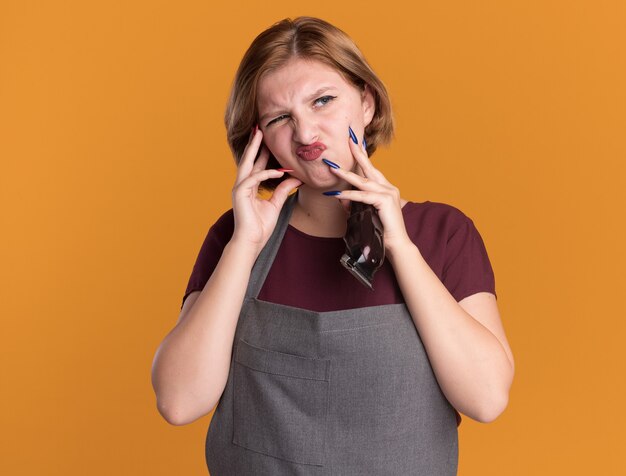 Dissatisfied young beautiful woman hairdresser in apron holding trimmer looking aside with confuse expression standing over orange wall