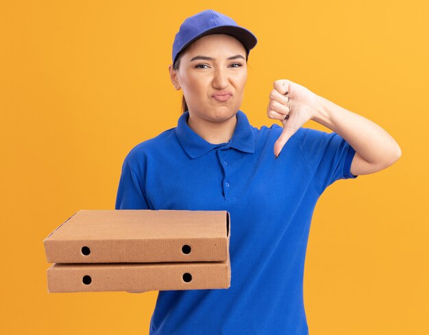 Dissatisfied young delivery woman in blue uniform and cap holding pizza boxes looking at front being displeased showing thumbs down standing over orange wall