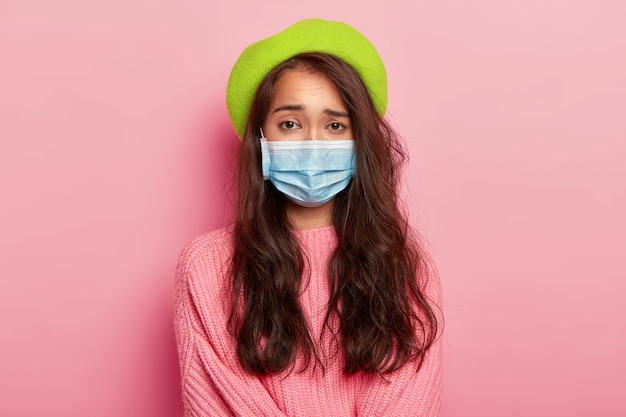 不満のある若い女性モデルは、医療用マスクを着用し、重病で、病院に来て医師の診察を受け、ウイルスに感染し、緑色のベレー帽とジャンパーを着用しています。