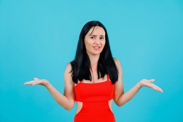 Dissatisfied young girl is looking at camera by opening wide her arms on blue background