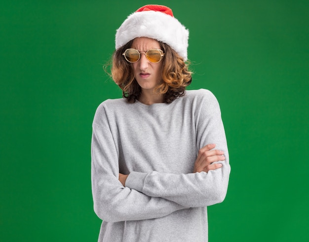 Dissatisfied young man wearing christmas santa hat and yellow glasses looking at camera with frowning face  with arms crossed standing over green background